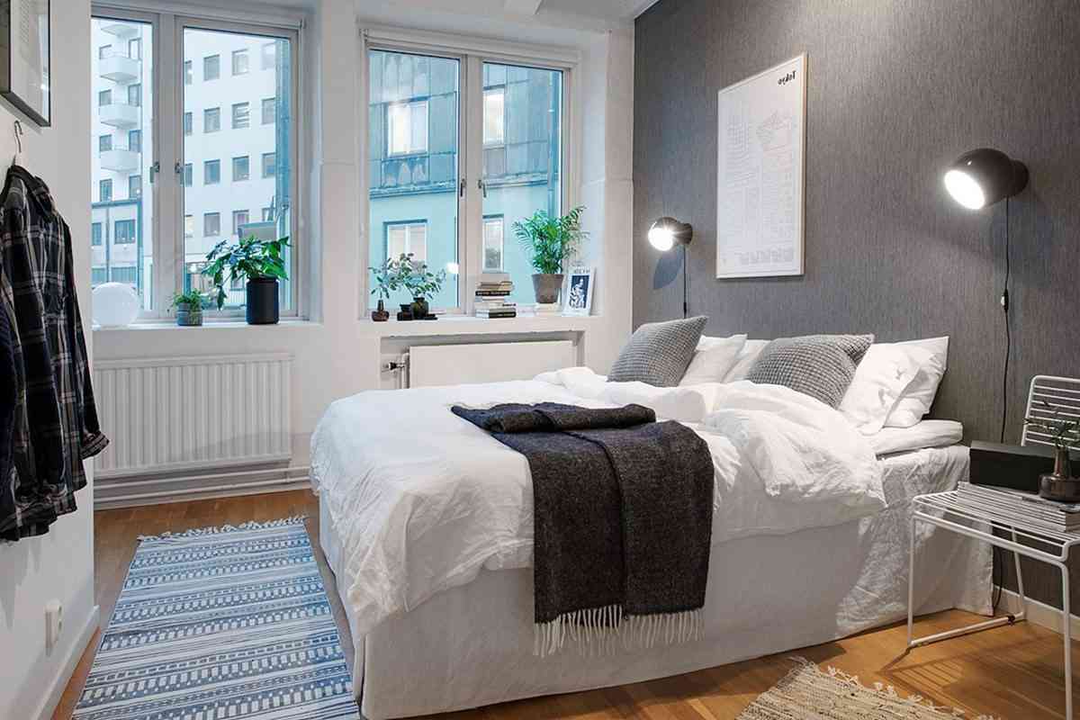 Ett modernt sovrum med en snyggt bäddad säng, grå vägg, hyllor med växter och ett fönster med utsikt över staden.